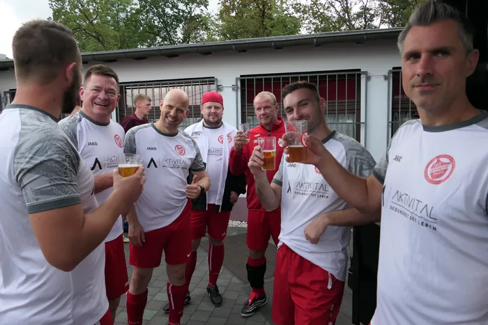 Eine Gruppe von Männern in weißen Sporttrikots und roten Hosen stößt mit Biergläsern an.