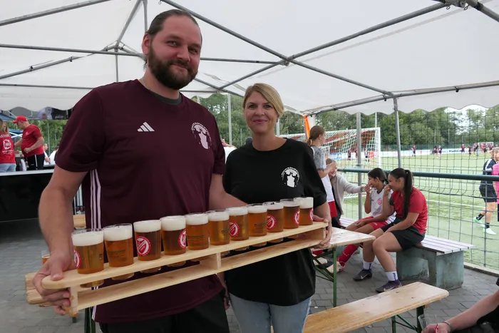 Ein Mann und eine Frau halten ein Holztablett mit vielen Biergläsern unter einem weißen Zelt bei einem Fußballturnier.