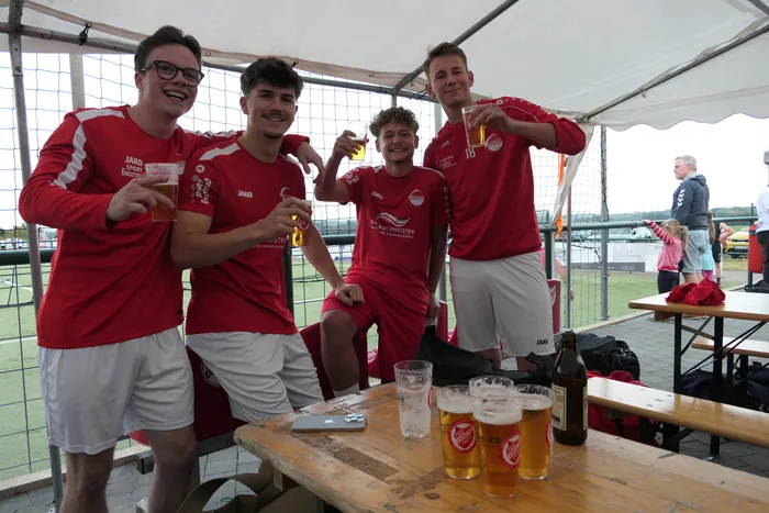 Vier junge Männer in roten Sporttrikots und weißen Shorts stoßen unter einem weißen Zelt mit Biergläsern an.