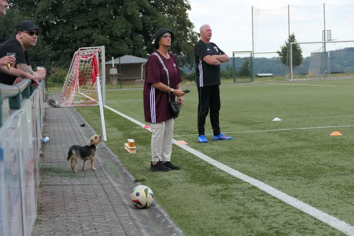 Eine Frau in weinrotem Trikot und ein Mann stehen am Spielfeldrand eines Fußballplatzes. Ein kleiner Hund und ein Fußball sind im Vordergrund.