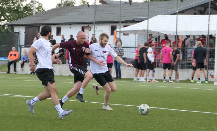 Drei Fußballspieler in weißen und weinroten Trikots kämpfen auf einem Rasenplatz um den Ball.