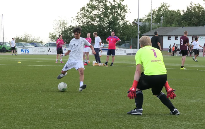 Ein Spieler in weißem Trikot dribbelt einen Fußball auf einem Kunstrasenplatz auf einen Torwart in einem neongelben Trikot zu.