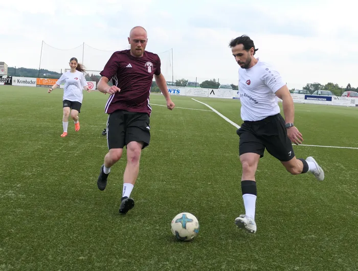Ein Mann in einem weinroten Trikot und ein Mann in einem weißen Trikot spielen um einen Fußball auf einem Kunstrasenplatz.