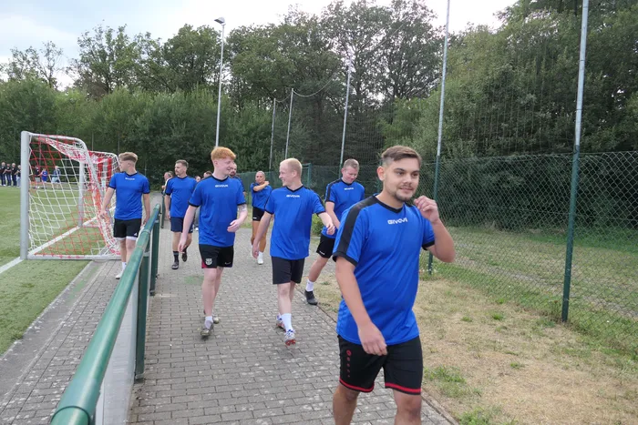 Mehrere junge Männer in blauen Fußballtrikots gehen auf einem gepflasterten Weg neben einem Fußballplatz.