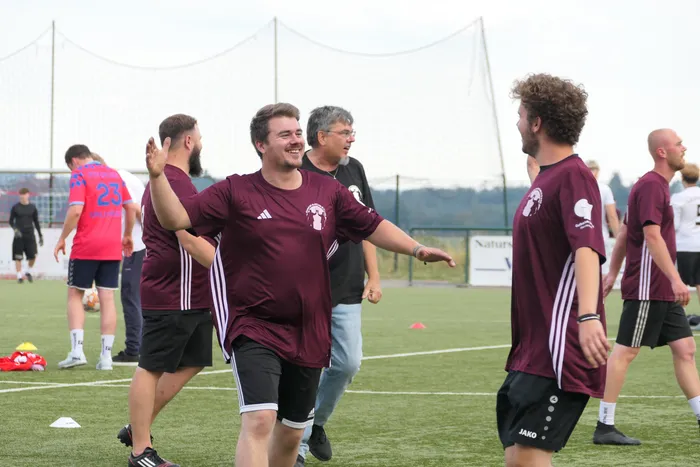 Ein lachender Mann in einem weinroten Trikot breitet die Arme aus, umgeben von anderen Spielern auf einem Fußballplatz.