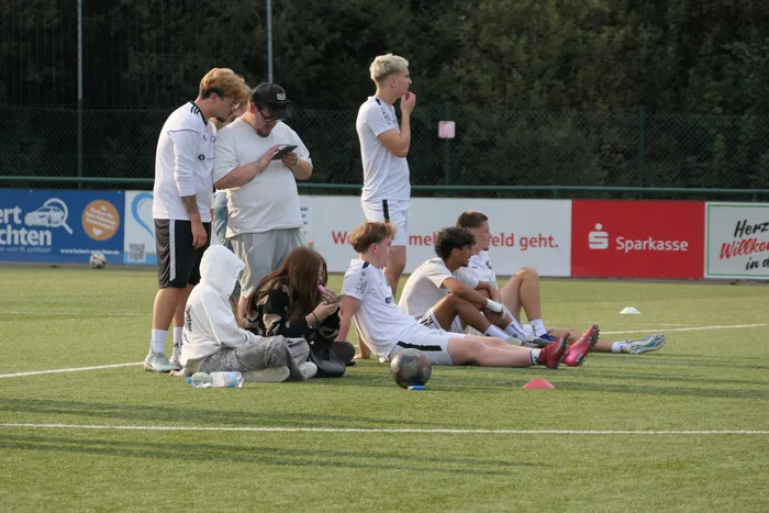 Eine Gruppe junger Menschen in weißer Sportkleidung sitzt und steht auf einem Fußballplatz während eines Hobby-Kicker-Turniers.