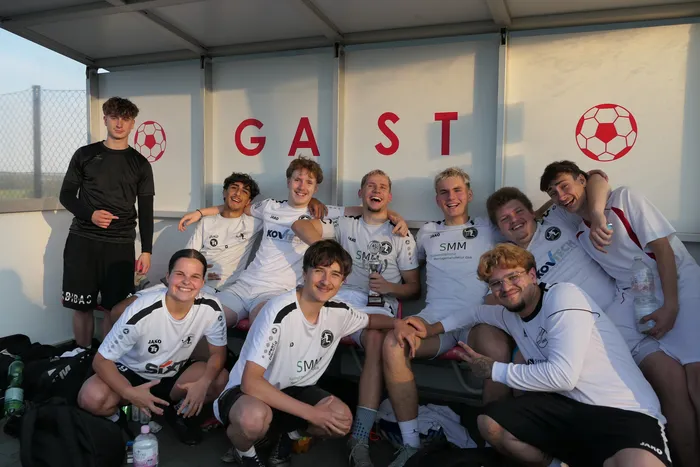 Eine Gruppe junger Fußballspieler in weißen Trikots posiert lächelnd vor einer „GAST“-Anzeige und hält einen Pokal.
