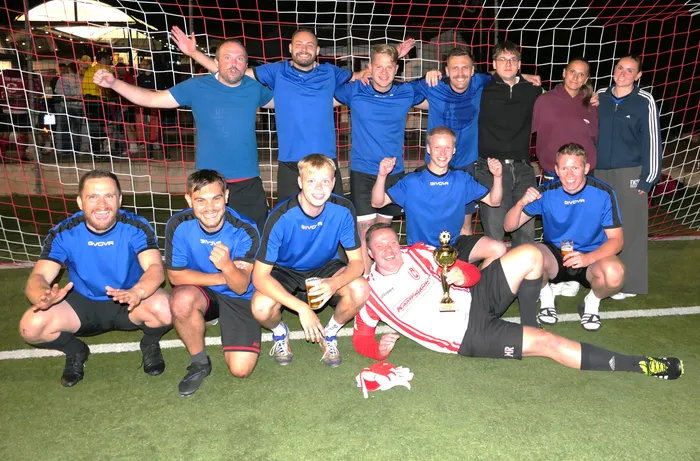 Eine Fußballmannschaft in blauen Trikots posiert vor einem Tornetz, ein Spieler in Rot-Weiß liegt mit einem Pokal auf dem Rasen.