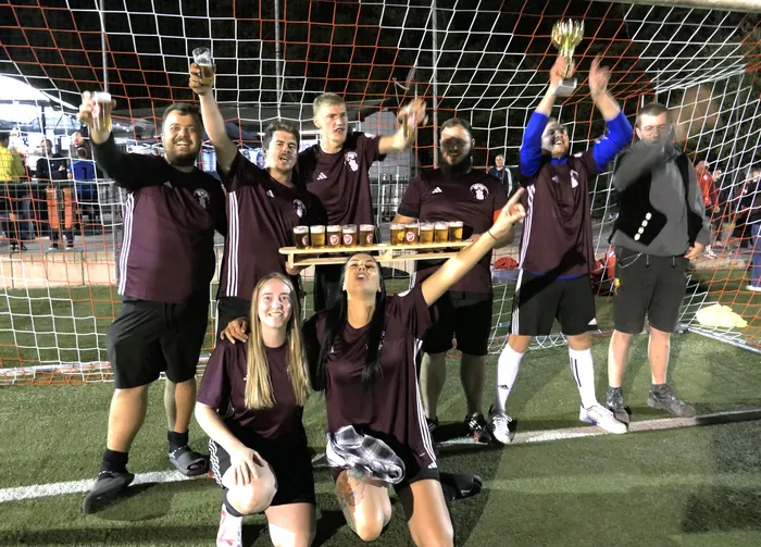 Eine Gruppe von Fußballspielern in weinroten Trikots feiert hinter einem Tornetz mit einem Pokal und Biergläsern.