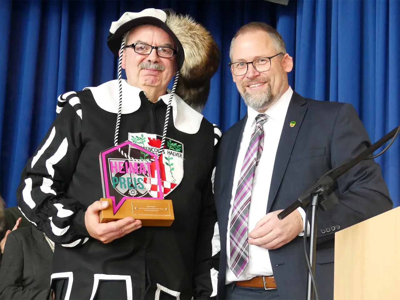 Ein Mann im schwarz-weißen Landsknecht-Kostüm hält die Auszeichnung „Heimatpreis“ neben einem Mann im dunklen Anzug.