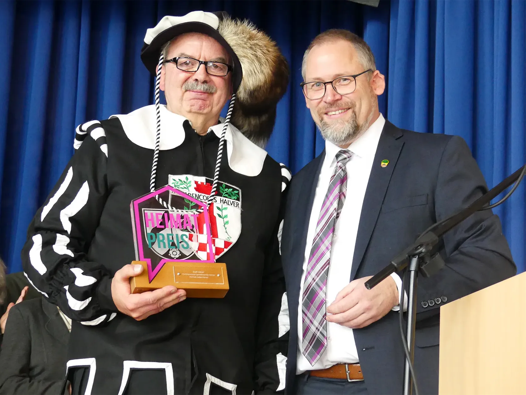 Ein Mann im schwarz-weißen Landsknecht-Kostüm hält die Auszeichnung „Heimatpreis“ neben einem Mann im dunklen Anzug.