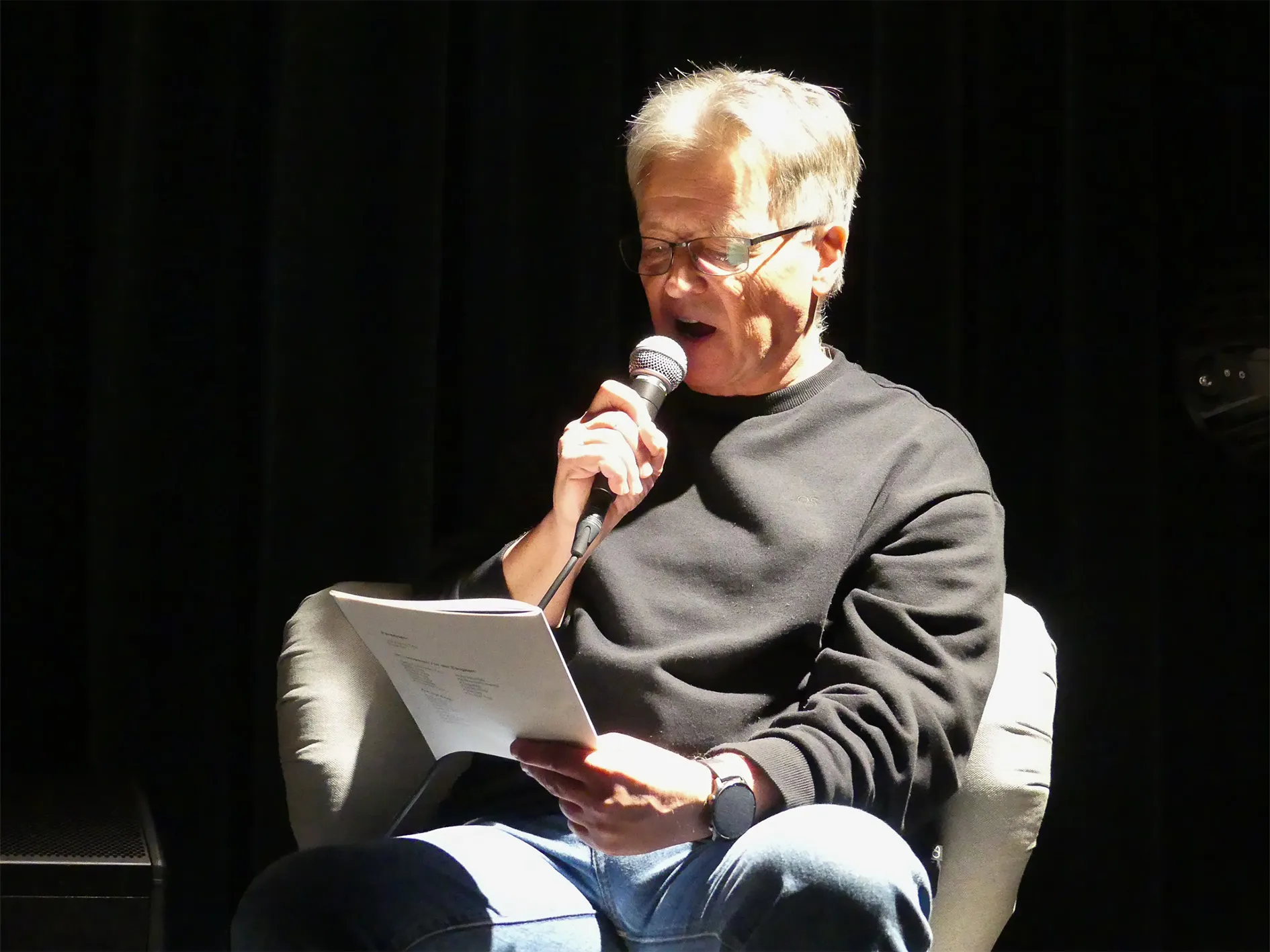 Ein älterer Mann mit Brille spricht in ein Mikrofon, während er auf ein Blatt Papier schaut. Er sitzt auf einem grauen Sessel und trägt Jeans und einen dunklen Pullover.