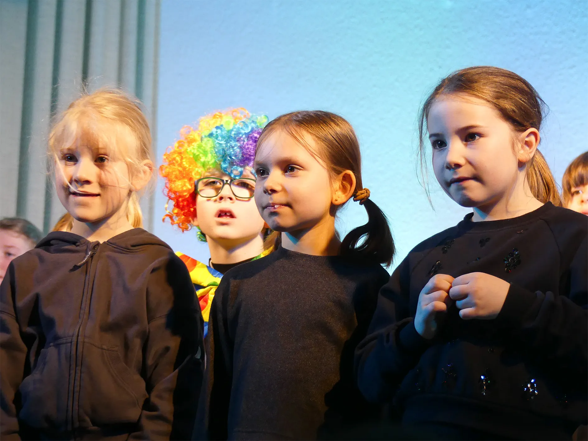Vier Kinder stehen auf einer Bühne, drei davon tragen dunkle Jacken. Im Hintergrund ist ein Kind mit bunter Lockenperücke und Brille zu sehen.
