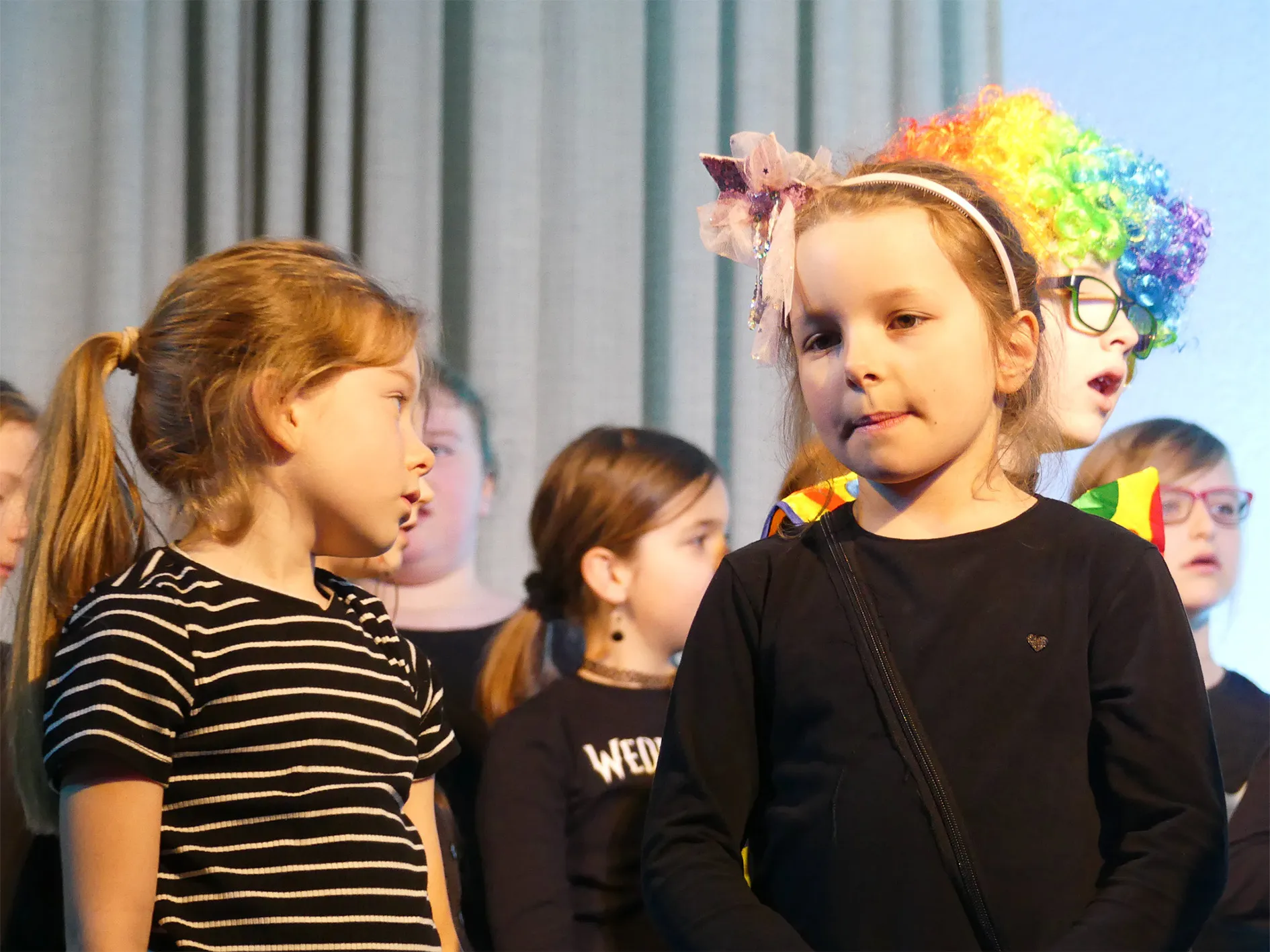Zwei Mädchen im Vordergrund während einer Theateraufführung, im Hintergrund weitere Kinder mit bunten Perücken und Kostümen.
