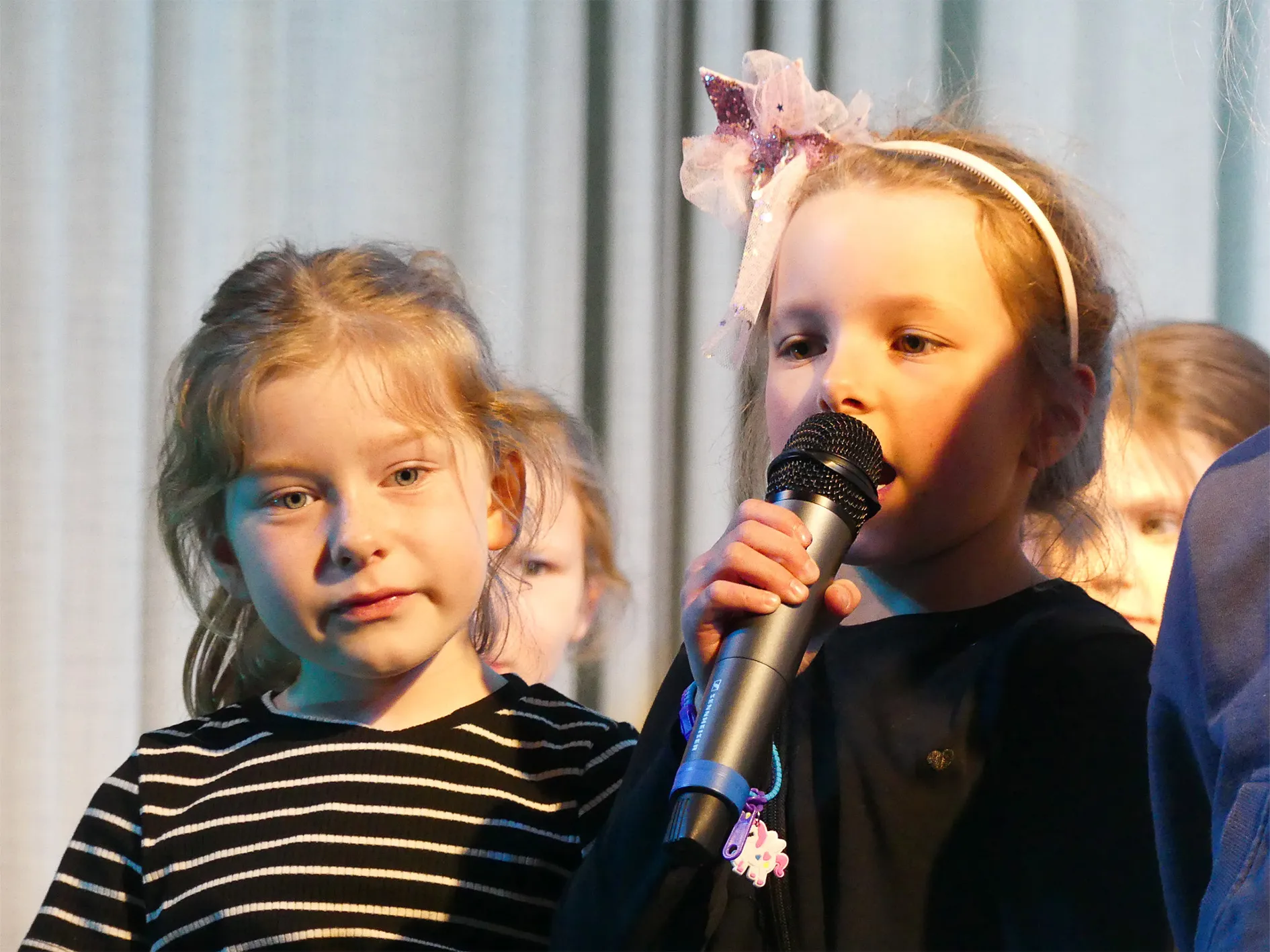 Zwei Mädchen singen auf einer Bühne, eines hält ein Mikrofon. Im Hintergrund sind weitere Kinder zu sehen.