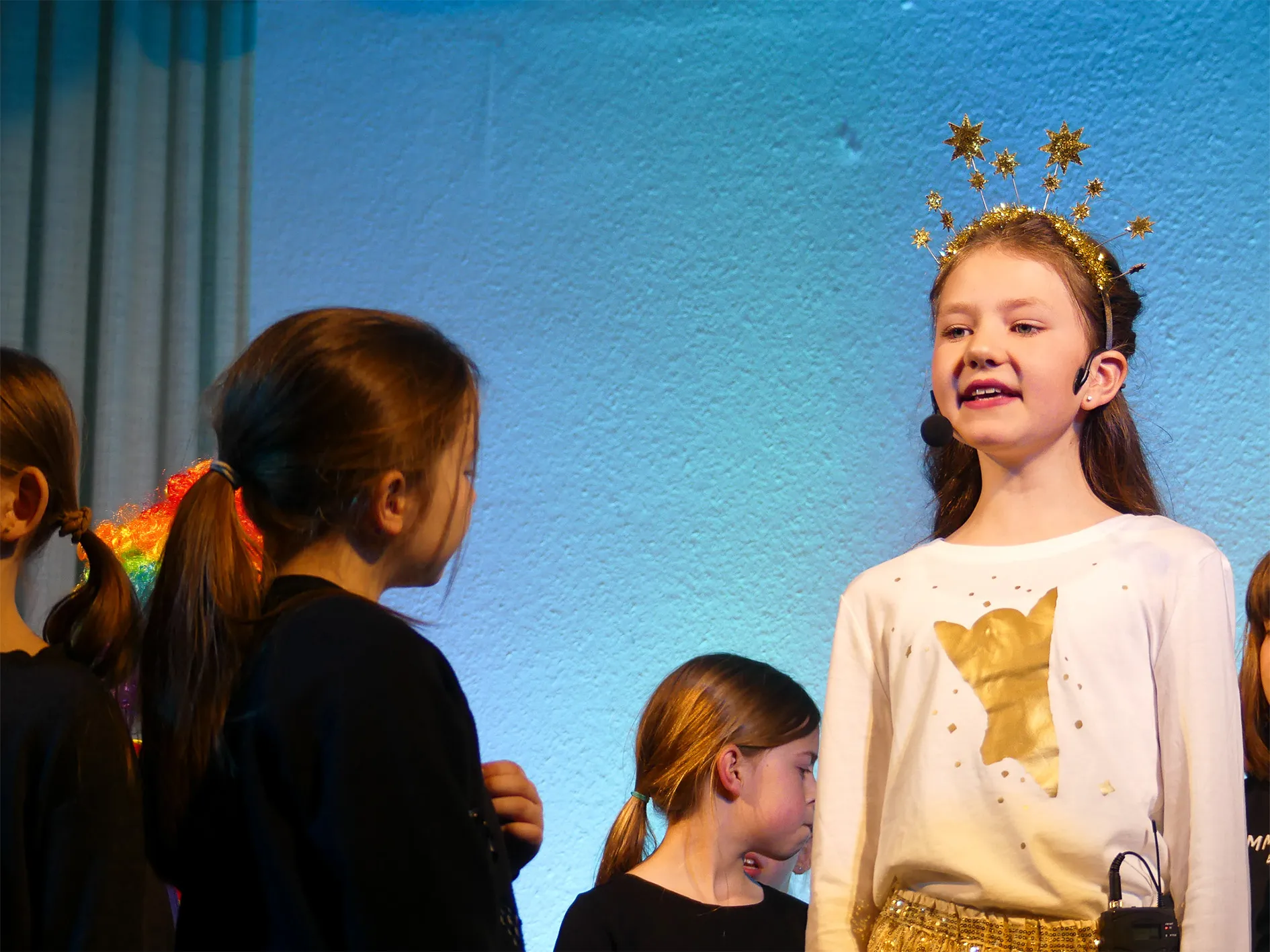 Ein Mädchen mit Sternenkrone singt in ein Headset, während andere Kinder der Grundschule Breckerfeld ihr zuhören.