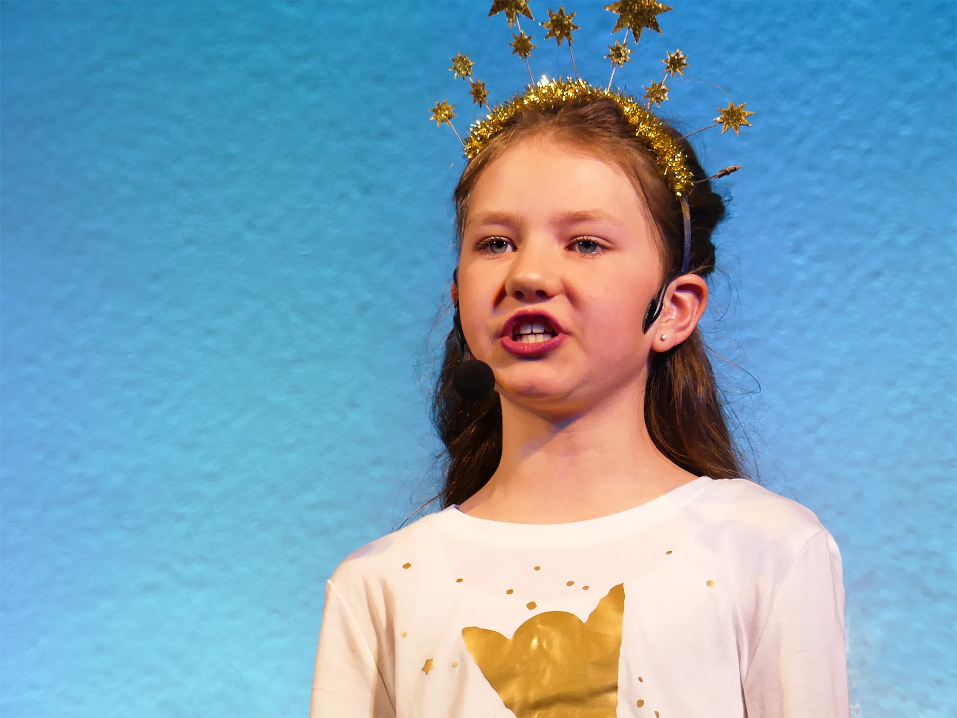 Ein Mädchen mit einem goldenen Sternenreif singt in ein Headset. Sie trägt ein weißes T-Shirt mit goldenem Aufdruck und steht auf einer Bühne vor blauem Hintergrund.
