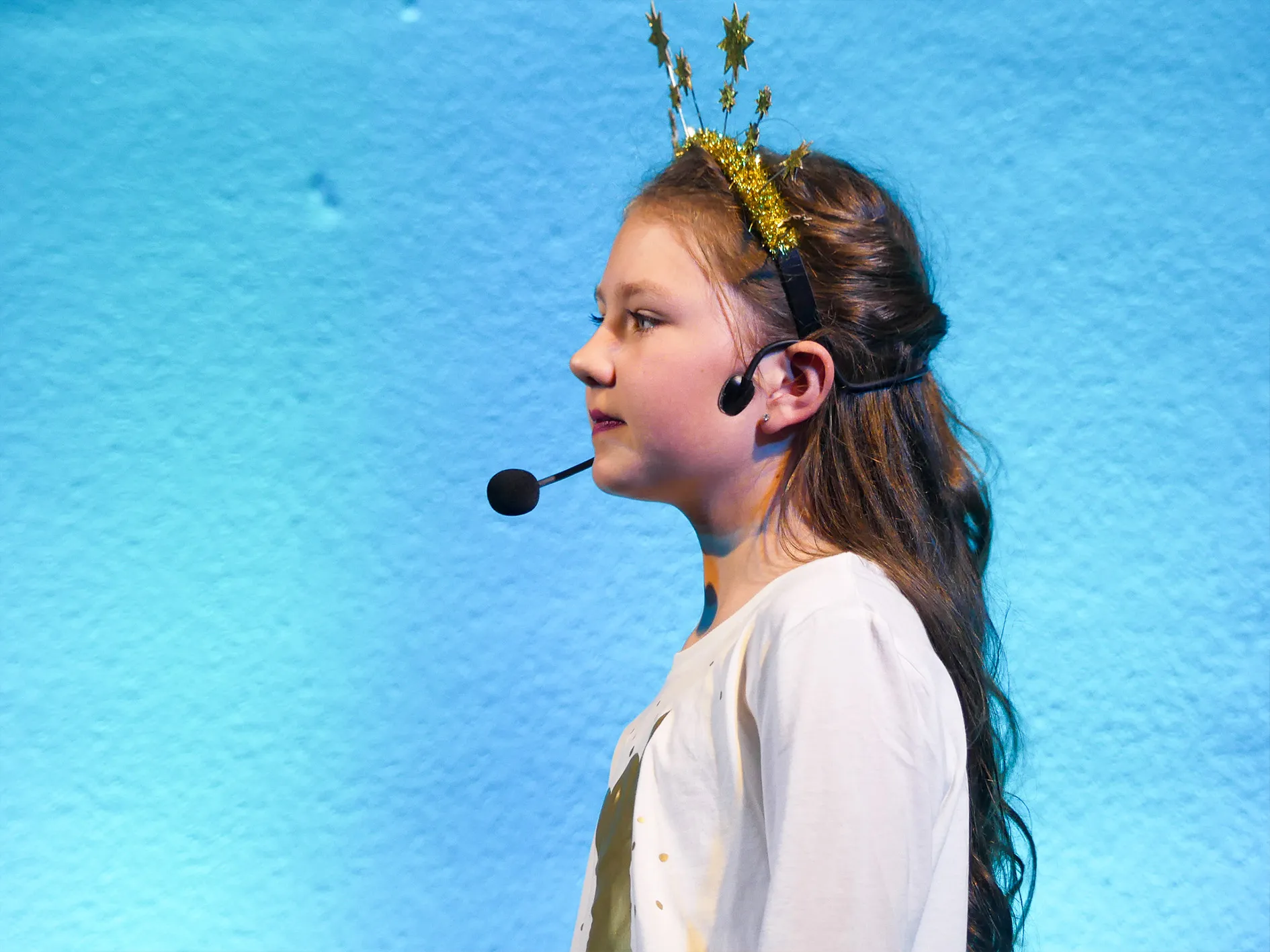 Ein Mädchen mit Stirnband und Mikrofon steht auf einer Bühne und trägt ein T-Shirt mit einem Aufdruck. Im Hintergrund eine blaue Wand.