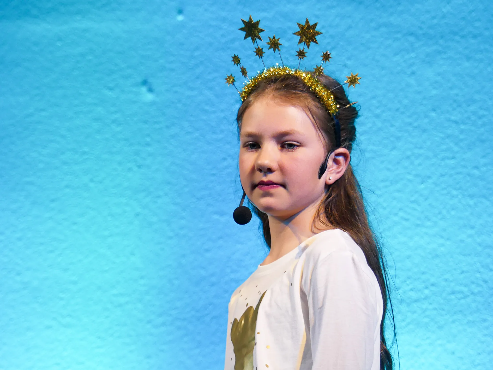 Ein Mädchen mit einem goldenen Sternen-Diadem und Headset steht auf einer Bühne und trägt ein weißes T-Shirt mit einem goldenen Aufdruck.