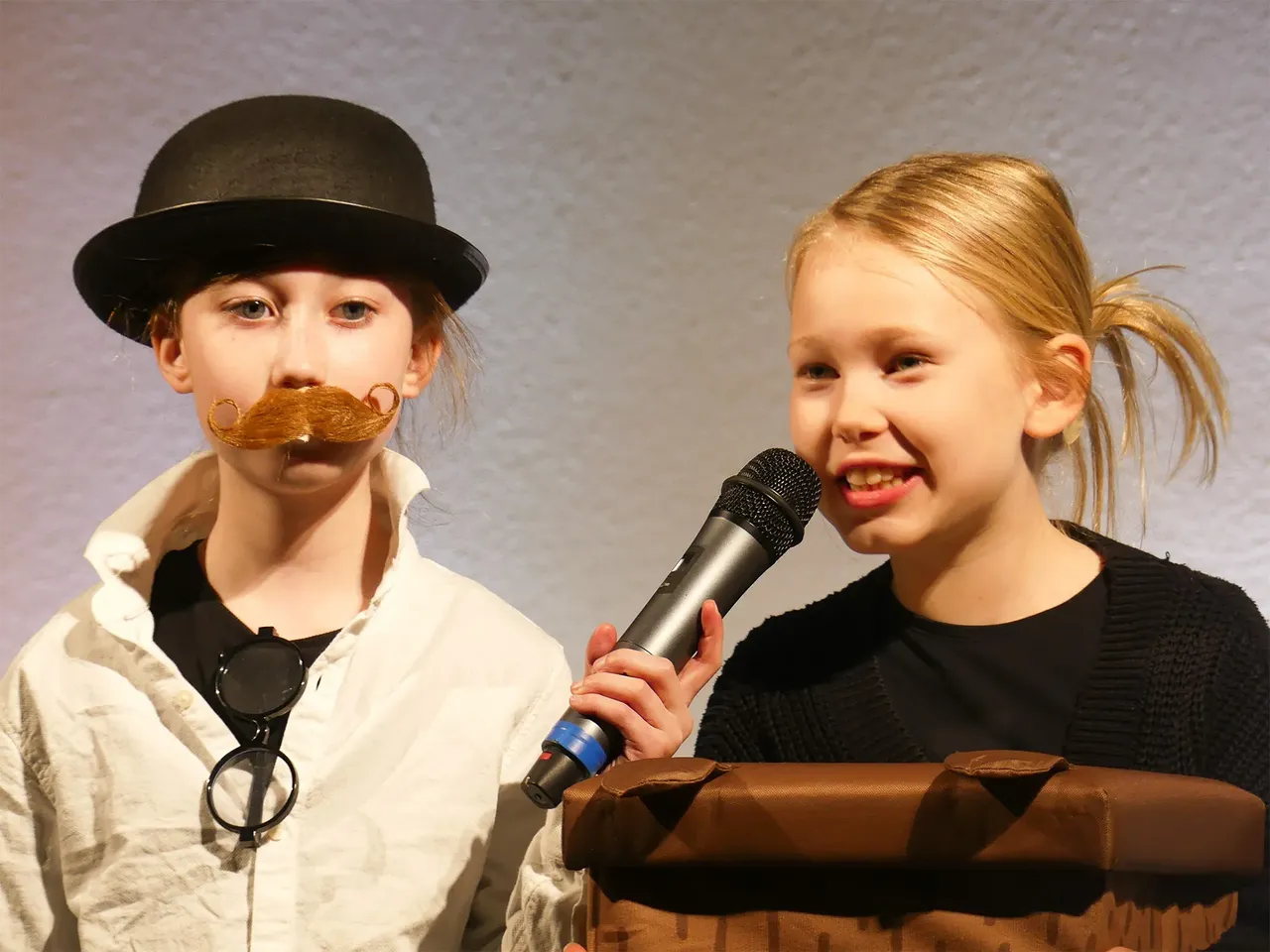 Zwei Kinder stehen auf einer Bühne und führen das Theaterstück „Der kleine Tag“ auf. Links trägt ein Kind einen Hut und einen Schnurrbart, rechts hält ein Kind ein Mikrofon und lächelt.