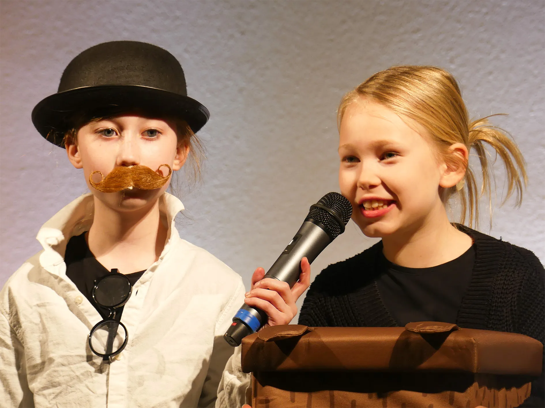 Zwei Kinder stehen auf einer Bühne und führen das Theaterstück „Der kleine Tag“ auf. Links trägt ein Kind einen Hut und einen Schnurrbart, rechts hält ein Kind ein Mikrofon und lächelt.