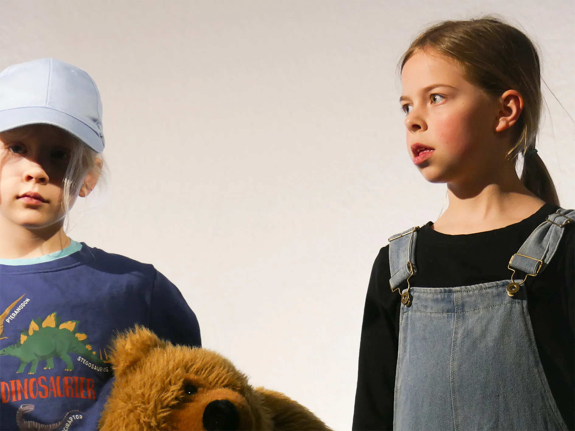 Zwei Kinder stehen nebeneinander vor einem weißen Hintergrund, das Mädchen blickt aufgeregt nach rechts, der Junge trägt eine Dinosaurier-Jacke und eine Kappe.