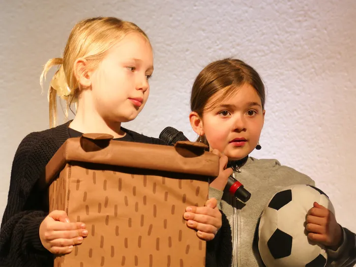 Zwei Mädchen halten Requisiten – ein Karton und einen Fußball – während einer Aufführung des Theaterstücks „Der kleine Tag“.