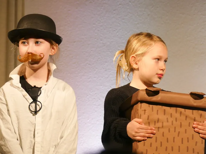 Zwei Kinder spielen eine Szene aus dem Theaterstück „Der kleine Tag“. Links trägt ein Kind einen Hut und einen Schnurrbart, rechts hält ein anderes Kind eine braune Pappkiste.