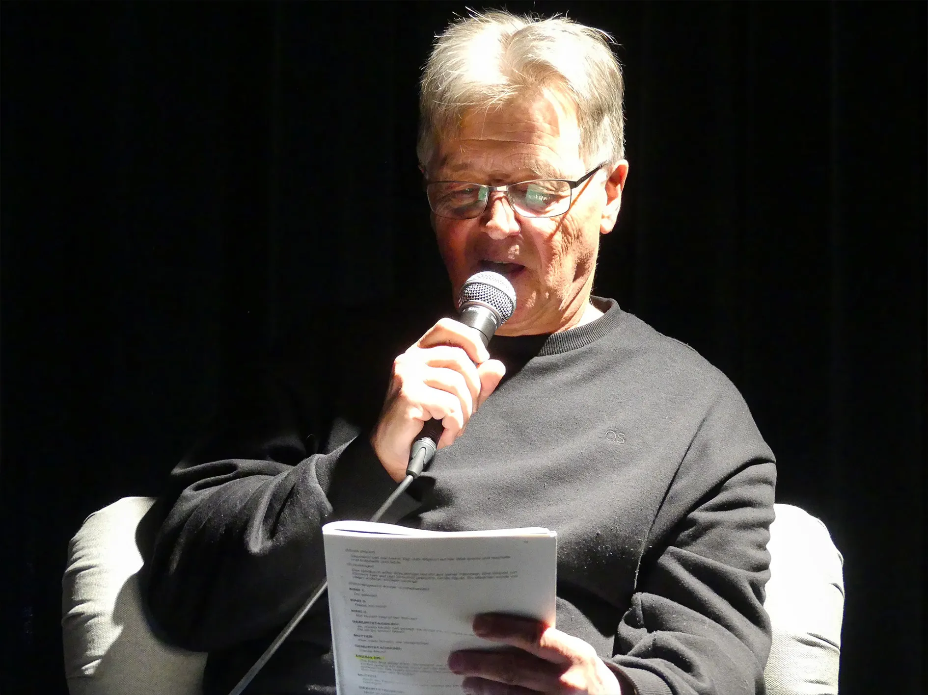 Ein älterer Mann mit grauen Haaren und Brille liest von einem Blatt Papier vor und hält ein Mikrofon in der Hand.