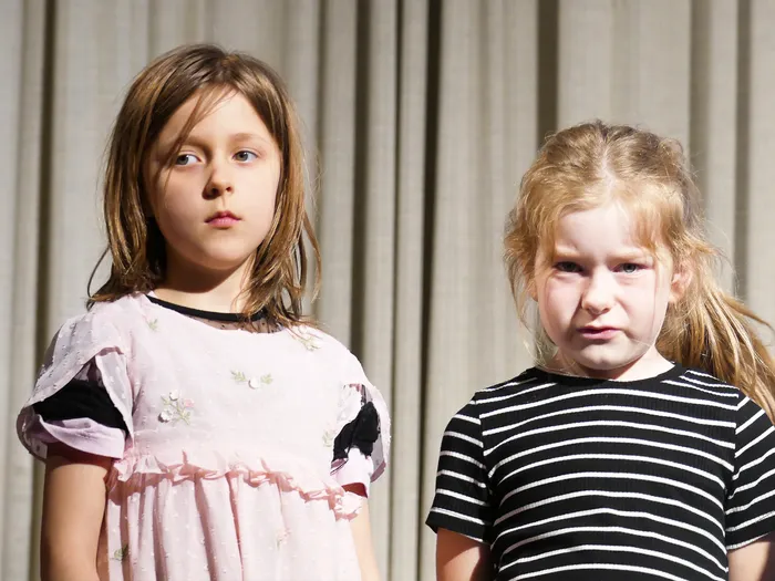 Zwei Mädchen im Schulalter stehen nebeneinander vor einem neutralen Hintergrund. Links trägt das Mädchen ein hellrosa Kleid, rechts ein schwarz-weiß gestreiftes Oberteil.