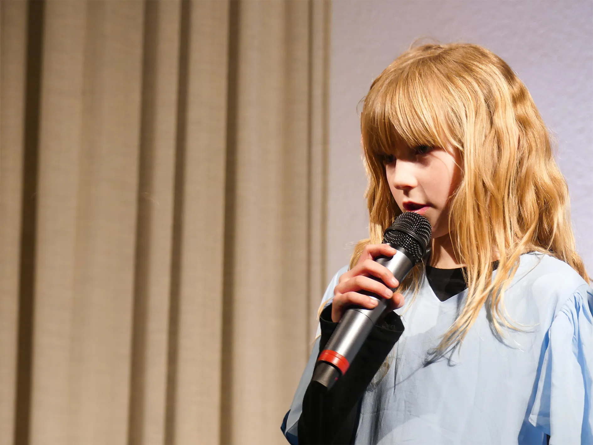 Ein blondes Kind mit Pony hält ein Mikrofon und spricht auf einer Bühne vor einem Vorhang. Das Kind trägt ein hellblaues Kleid.
