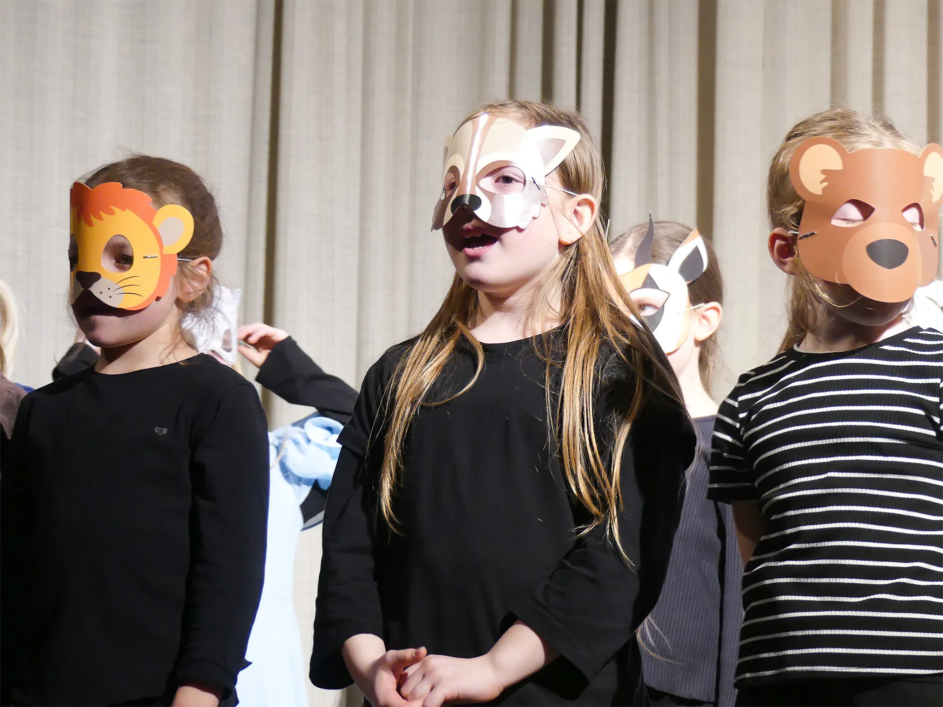 Kinder mit Tiermasken – Löwe, Fuchs und Bär – stehen auf einer Bühne und singen oder sprechen.