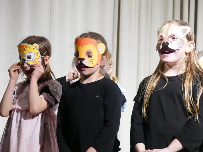Drei Kinder tragen Tiergesichtsmasken und scheinen ein Theaterstück aufzuführen. Links eine Maske mit Fuchsgesicht, in der Mitte ein Löwe und rechts ein Hirsch.
