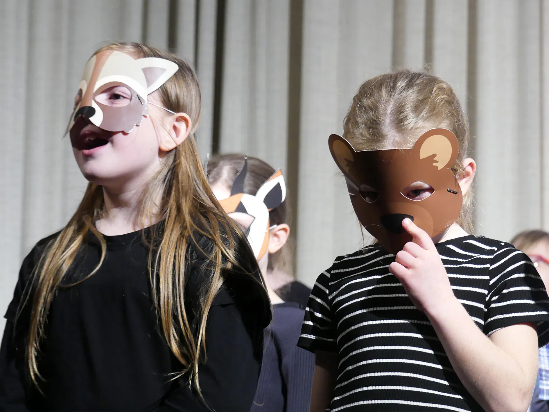 Zwei Mädchen mit Tiermasken im Vordergrund, weitere Kinder mit Masken im Hintergrund während einer Aufführung.