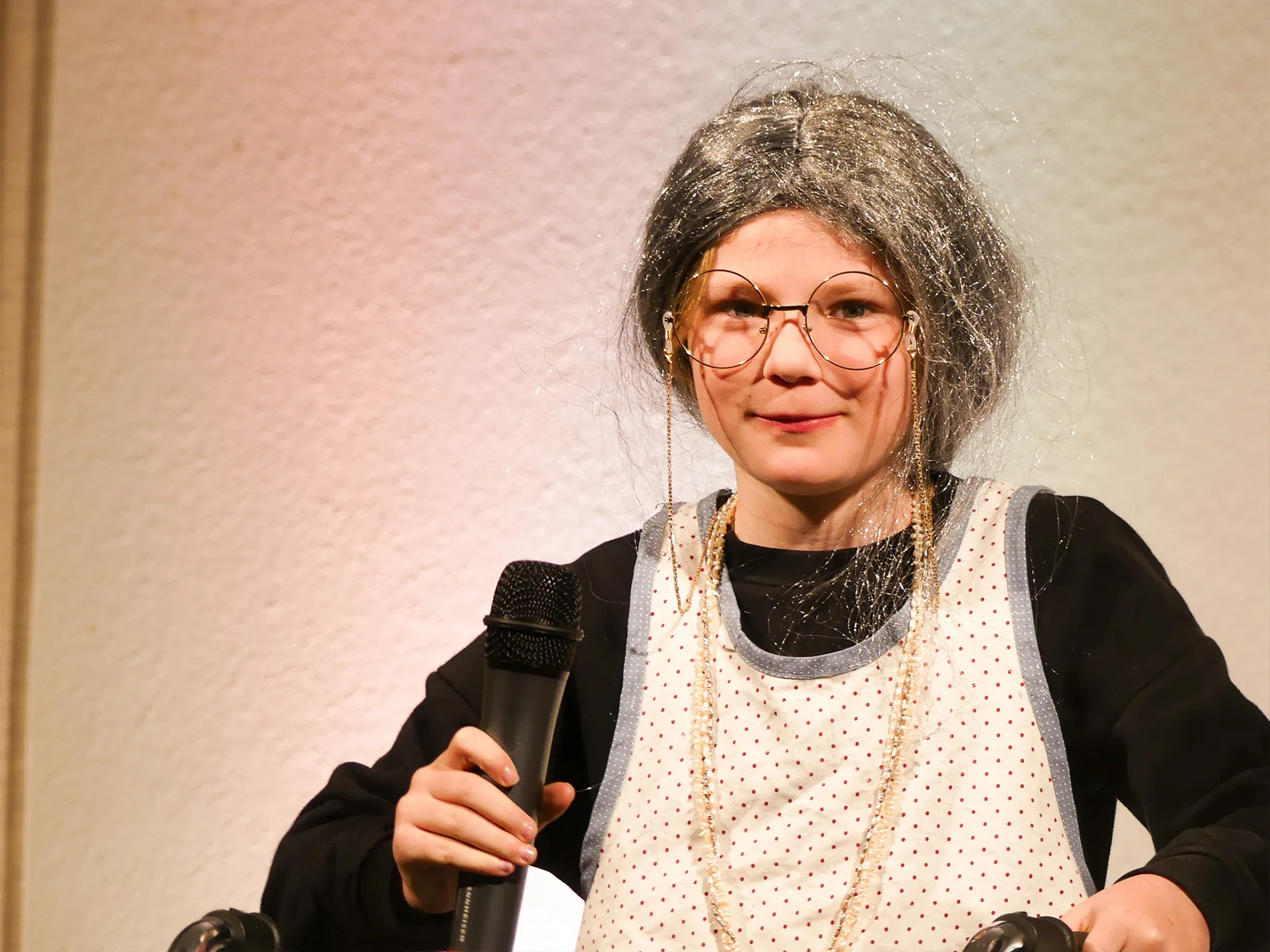 Frau mit grauer Perücke, Brille und roter Weste hält ein Mikrofon und spricht.