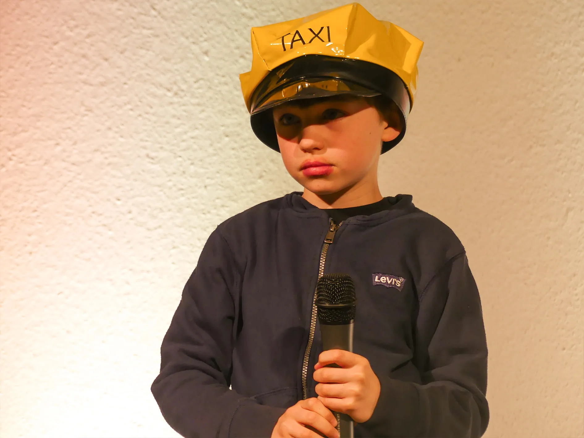 Junge mit Taxi-Mütze hält Mikrofon. Er trägt eine dunkle Jacke und steht vor einer hellen Wand.