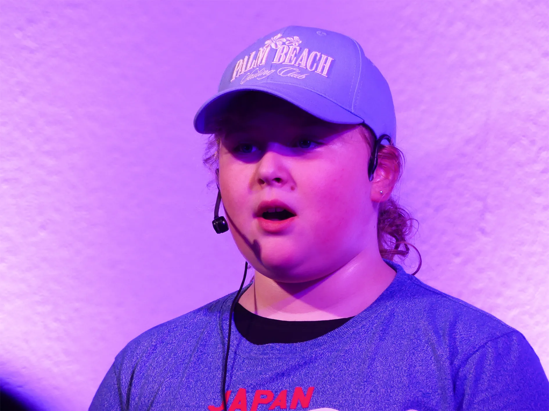 Ein Kind mit Headset singt auf der Bühne, trägt eine Baseballkappe mit dem Aufdruck „Palm Beach“ und ein T-Shirt mit dem Schriftzug „Japan“.
