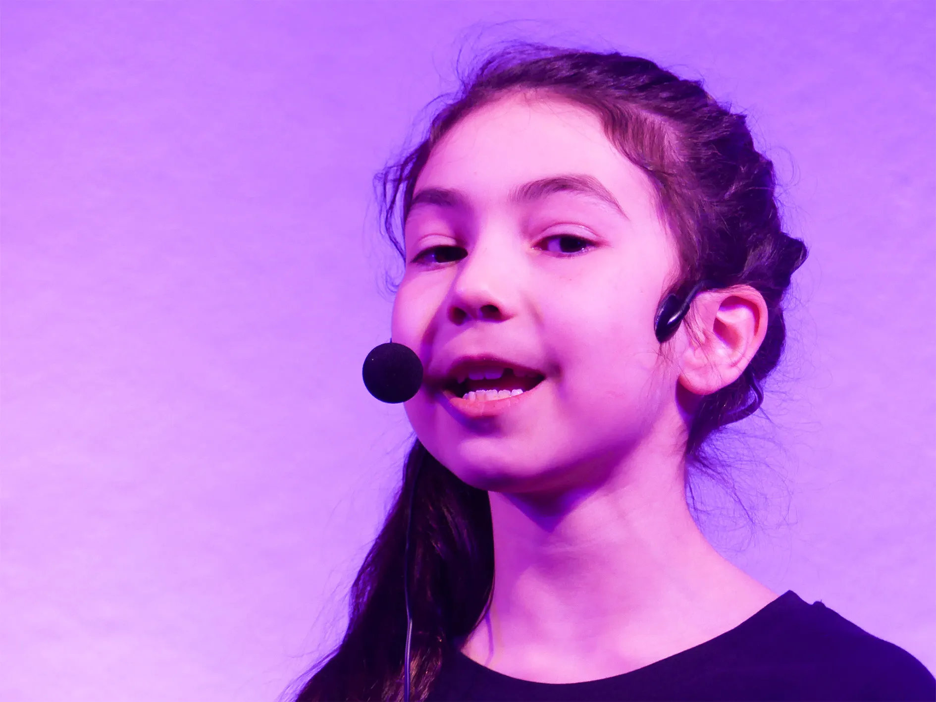 Ein Mädchen mit dunklem, gewelltem Haar spricht in ein Mikrofon. Im Hintergrund ist lila Licht zu sehen.
