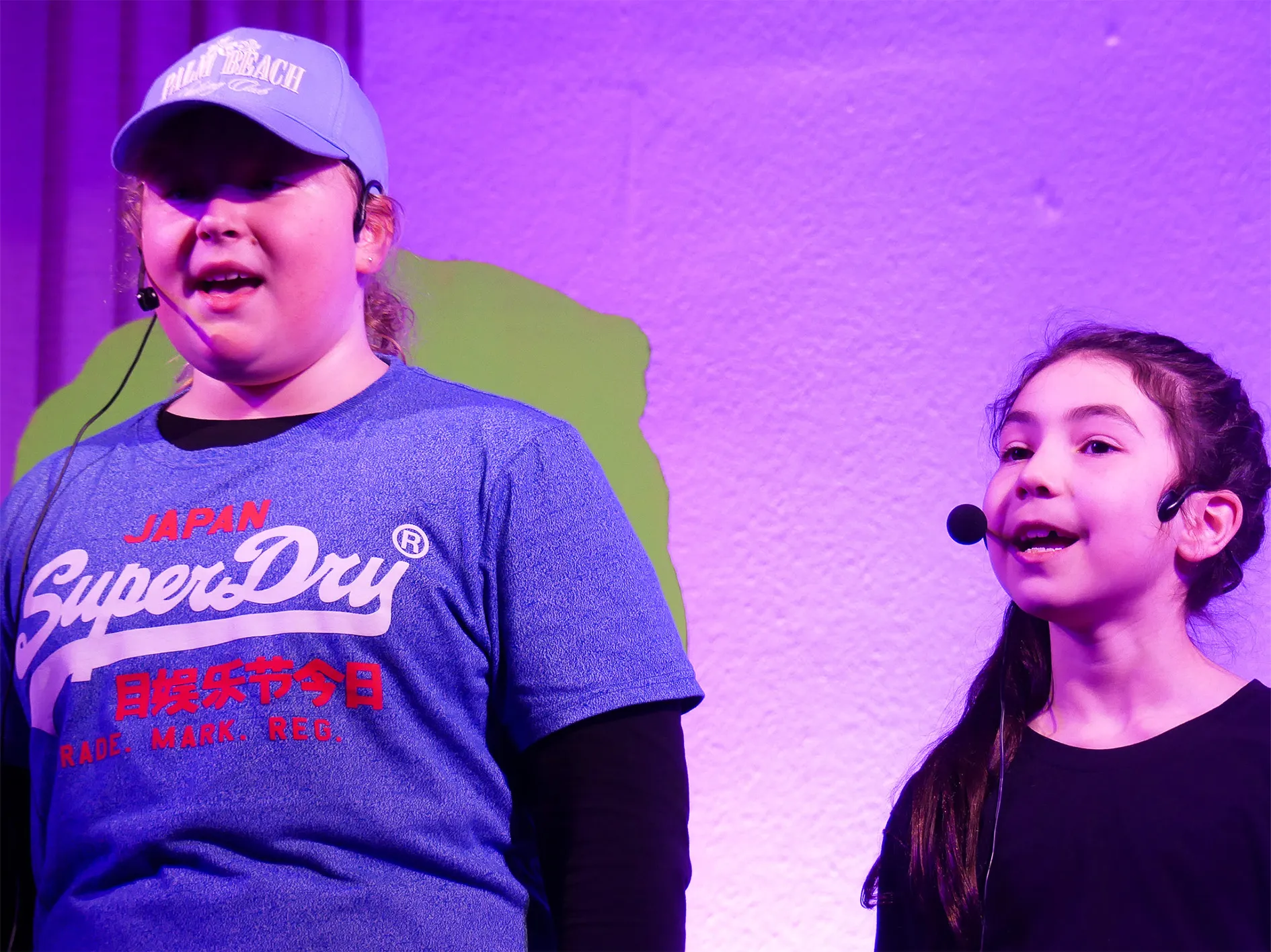 Zwei Kinder stehen auf einer Bühne und sprechen mit Headsets ins Publikum. Der Junge trägt eine blaue Jacke mit Aufdruck und eine Baseballkappe, das Mädchen ein rosa Oberteil.