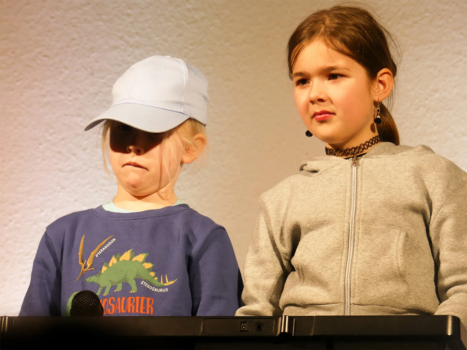 Zwei Kinder stehen auf einer Bühne, beide tragen Headsets. Der Junge links trägt eine Baseballkappe und ein T-Shirt mit einem Dinosaurier-Motiv, das Mädchen rechts eine graue Jacke.