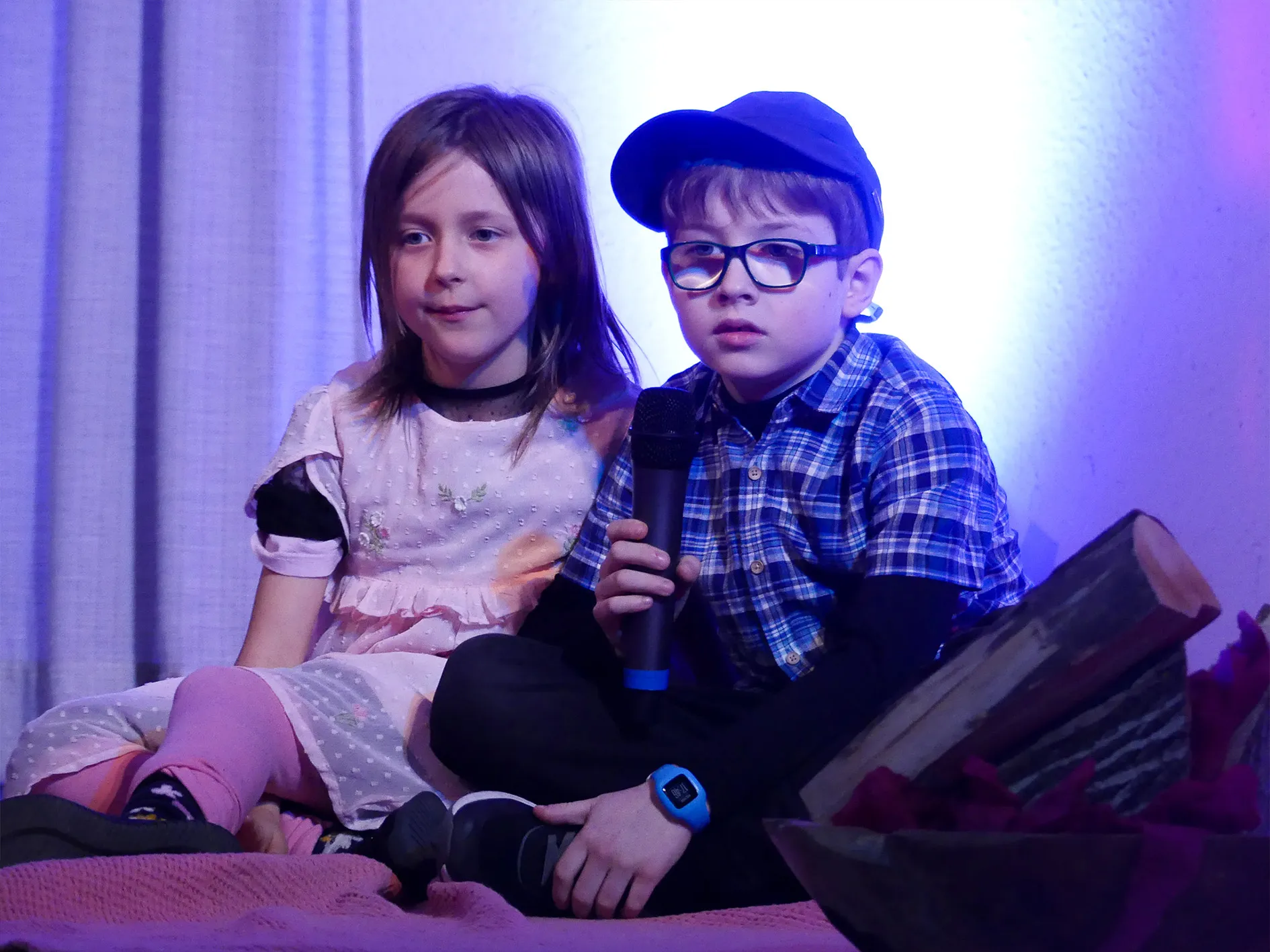 Zwei Kinder sitzen nebeneinander auf einer Couch, ein Junge mit Brille hält ein Mikrofon. Im Hintergrund ist ein Fenster mit Vorhängen zu sehen.