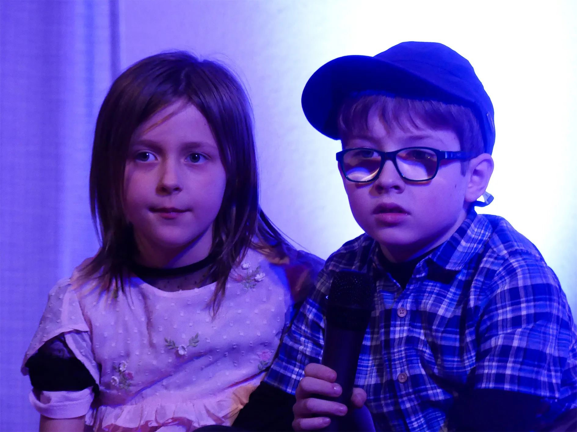 Zwei Kinder, ein Mädchen und ein Junge, blicken zur Kamera. Der Junge hält ein Mikrofon und trägt eine Mütze und Brille.