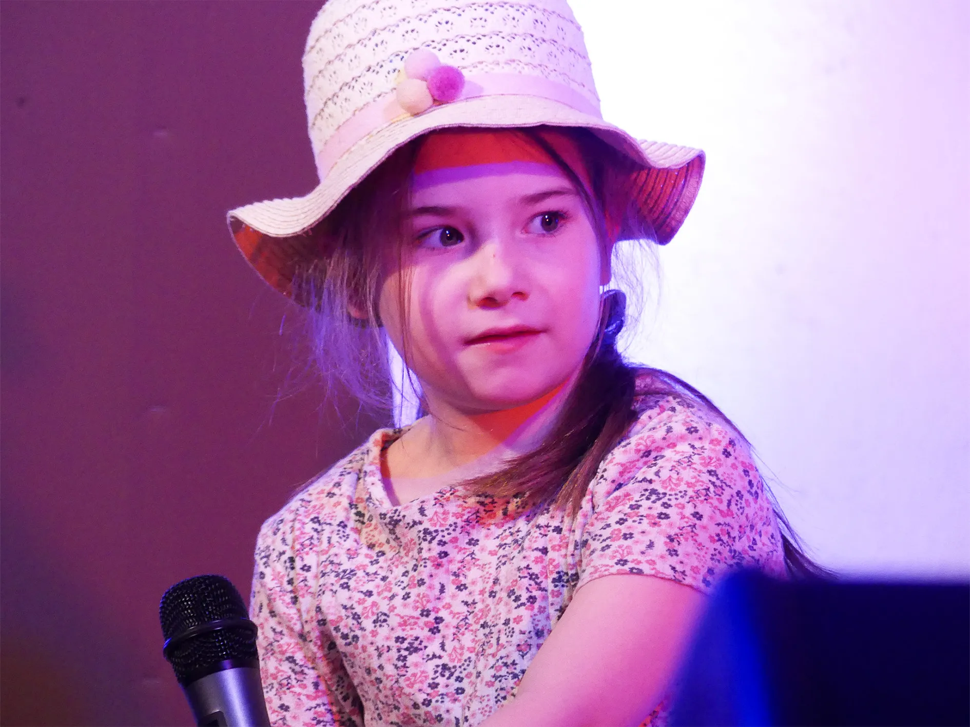 Jüngeres Mädchen mit Down-Syndrom trägt einen Strohhut und ein geblümtes Kleid, während sie in einer Bühnenaufführung spricht.