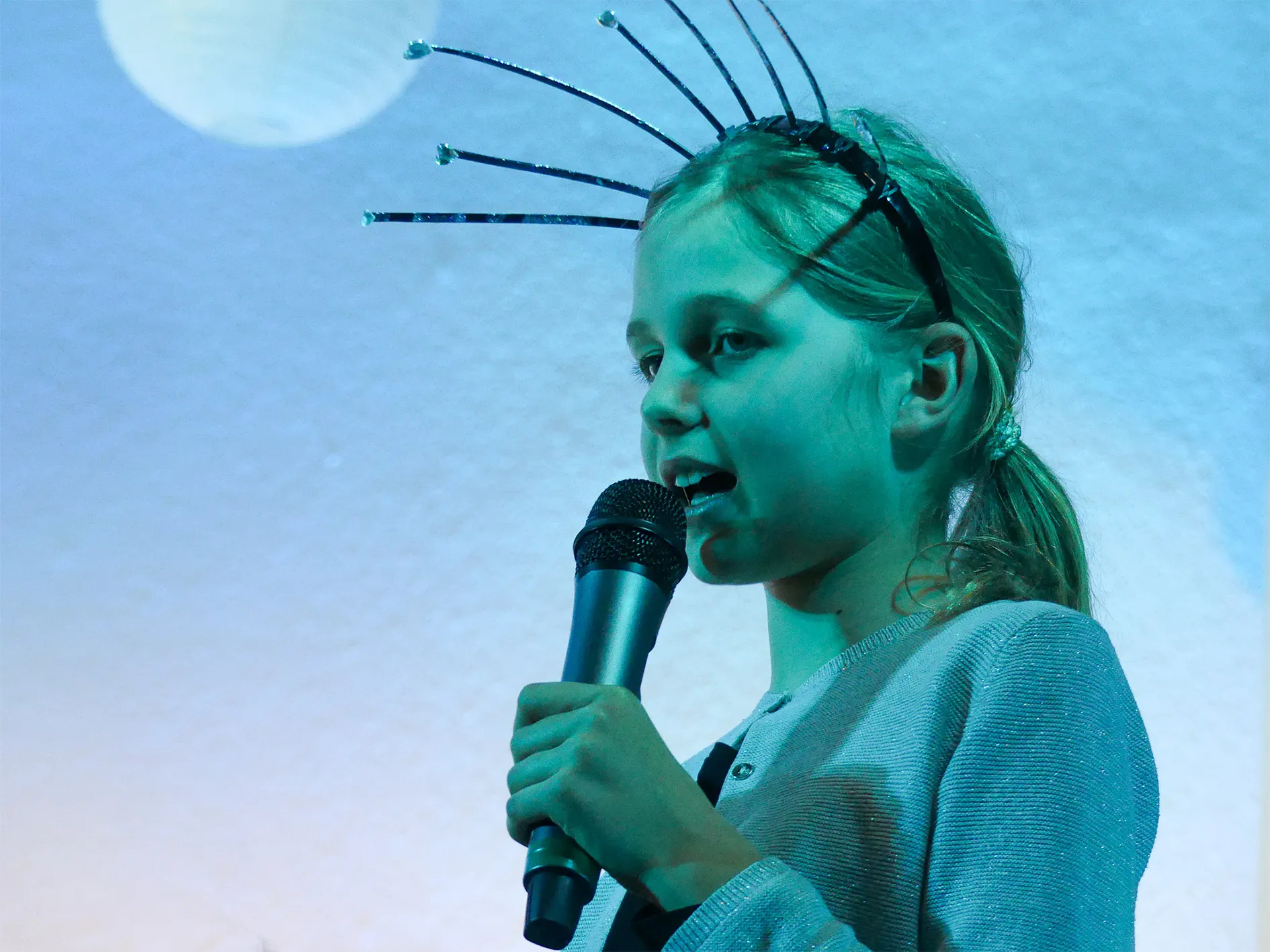 Jüngeres Mädchen mit Antennen-Haarreif spricht in ein Mikrofon, im Hintergrund eine stilisierte Mondkulisse.