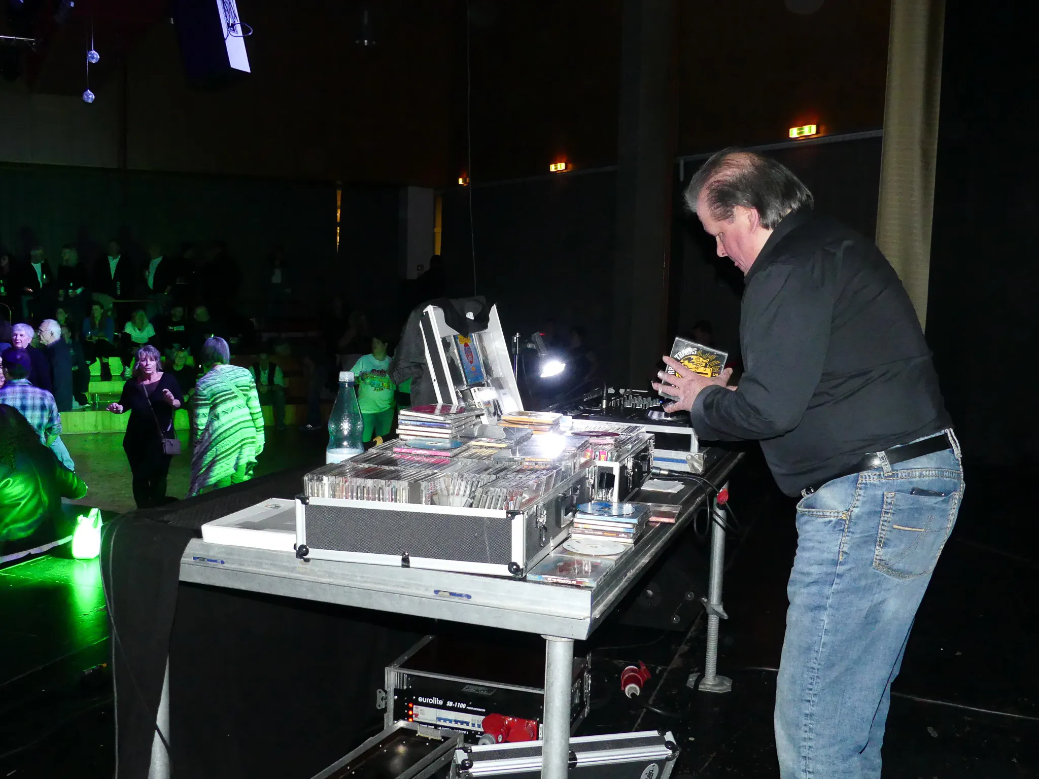 Ein DJ steht hinter einem Tisch voller CDs und bedient die Geräte. Im Hintergrund tanzen Menschen in einem abgedunkelten Raum.