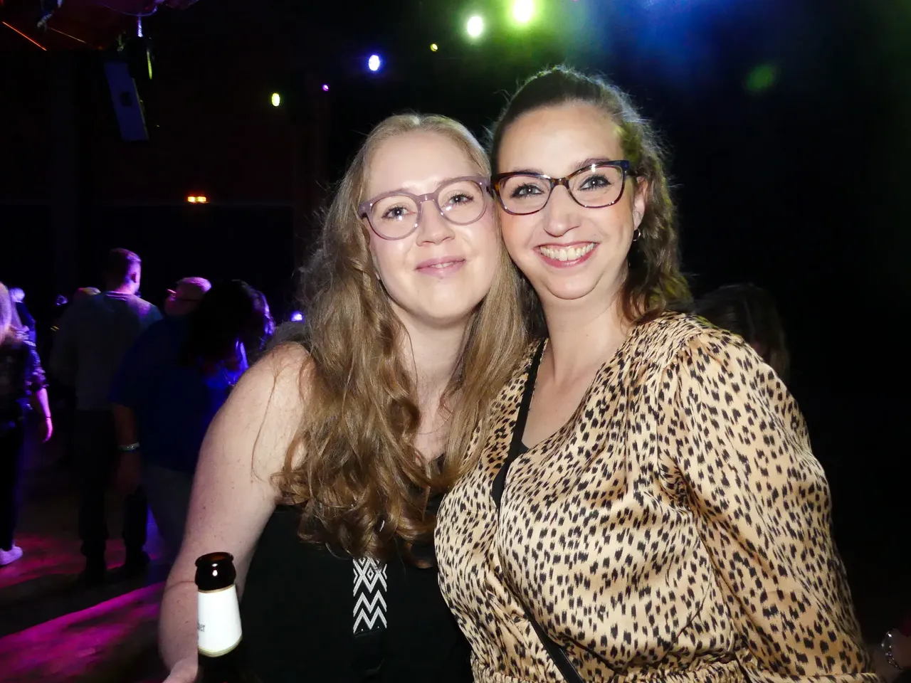 Zwei junge Frauen mit Brille lächeln in die Kamera, im Hintergrund eine tanzende Menschenmenge in einem dunklen Raum mit Bühnenbeleuchtung.