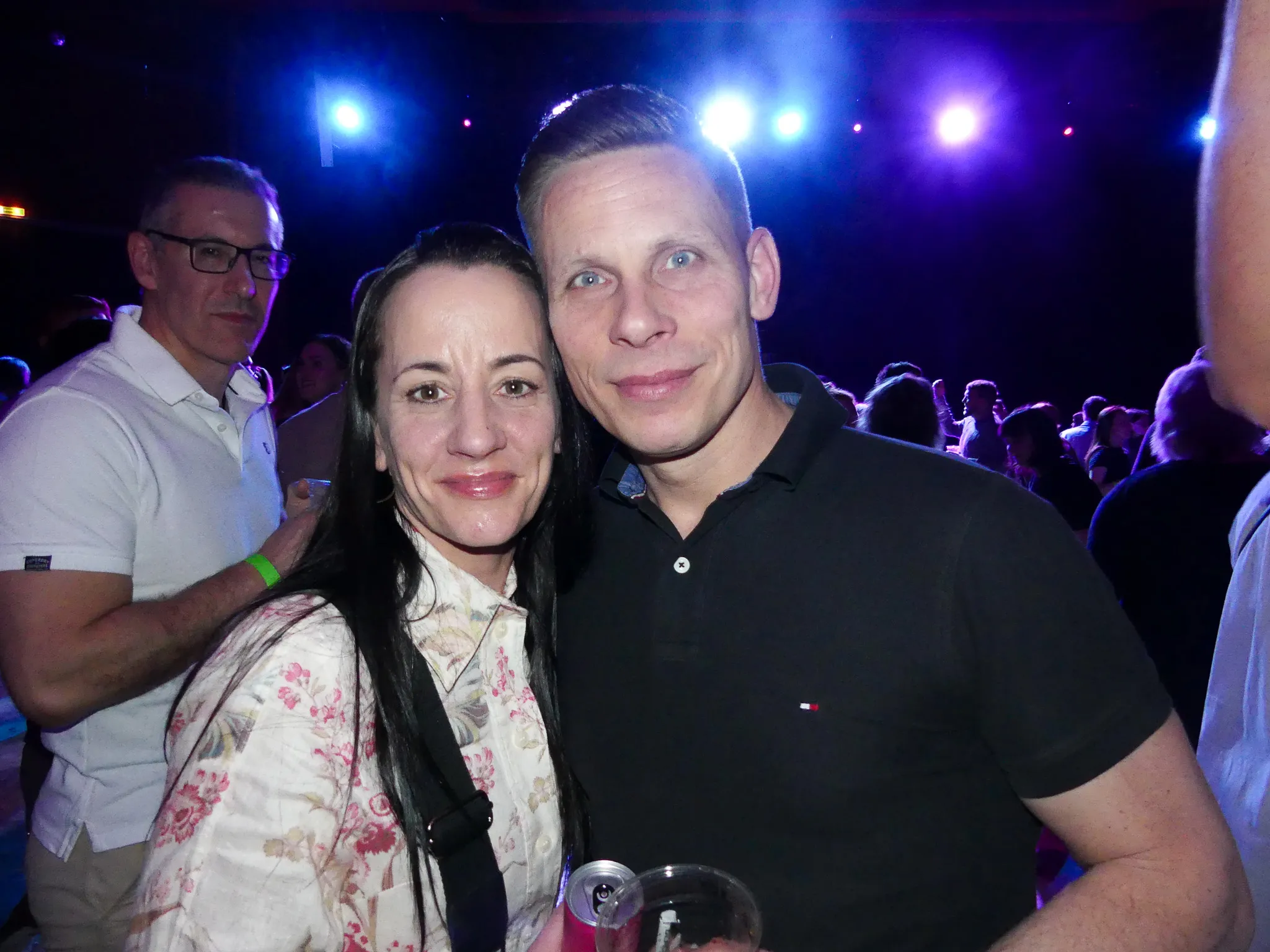 Ein Paar lächelt in die Kamera, umgeben von Menschen auf einer Tanzfläche mit bunter Beleuchtung.