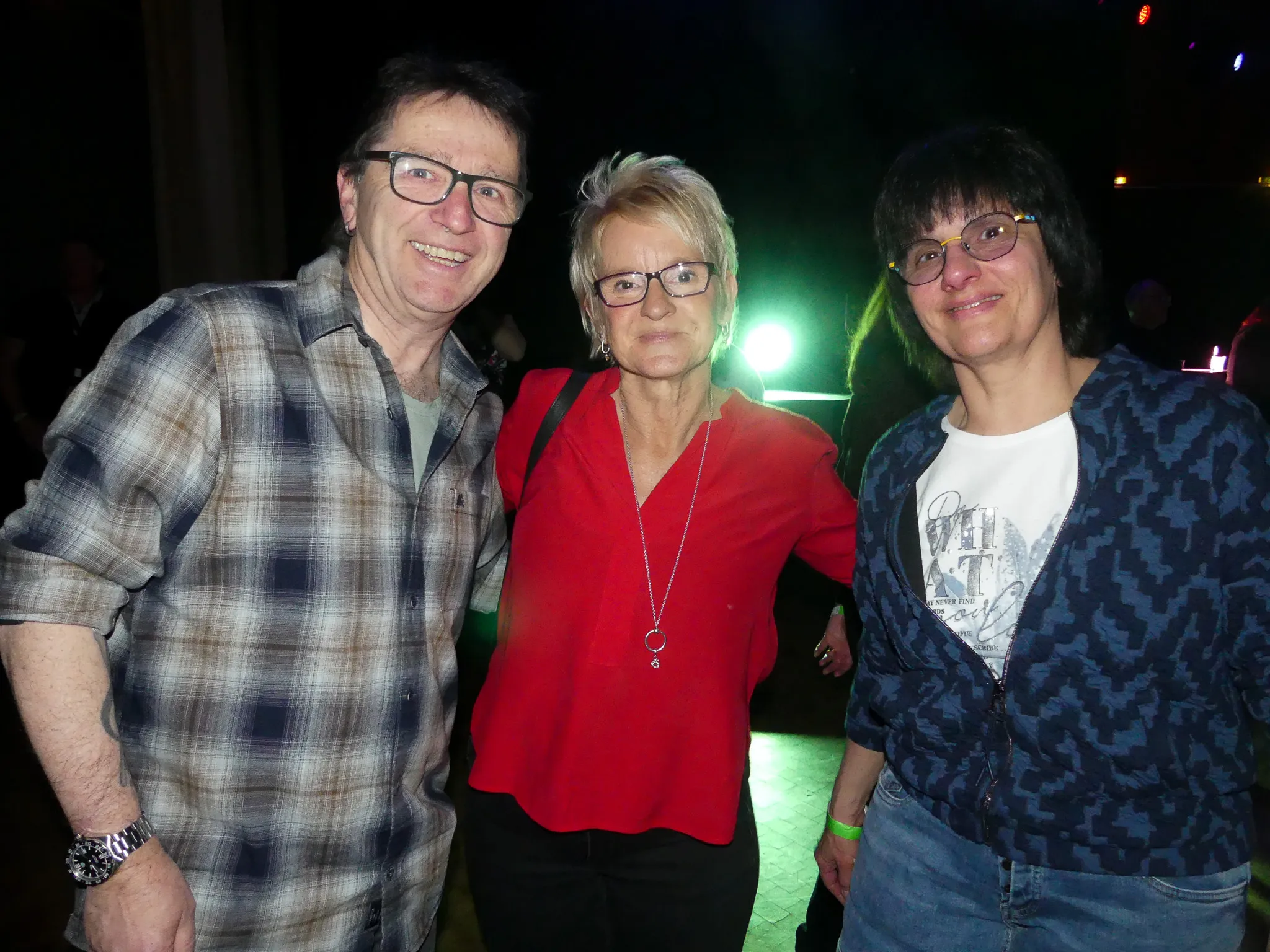 Drei lächelnde Menschen stehen eng zusammen, vermutlich auf einer Party. Links ein Mann mit kariertem Hemd und Brille, in der Mitte eine Frau in rotem Oberteil und rechts eine Frau mit bedrucktem T-Shirt.