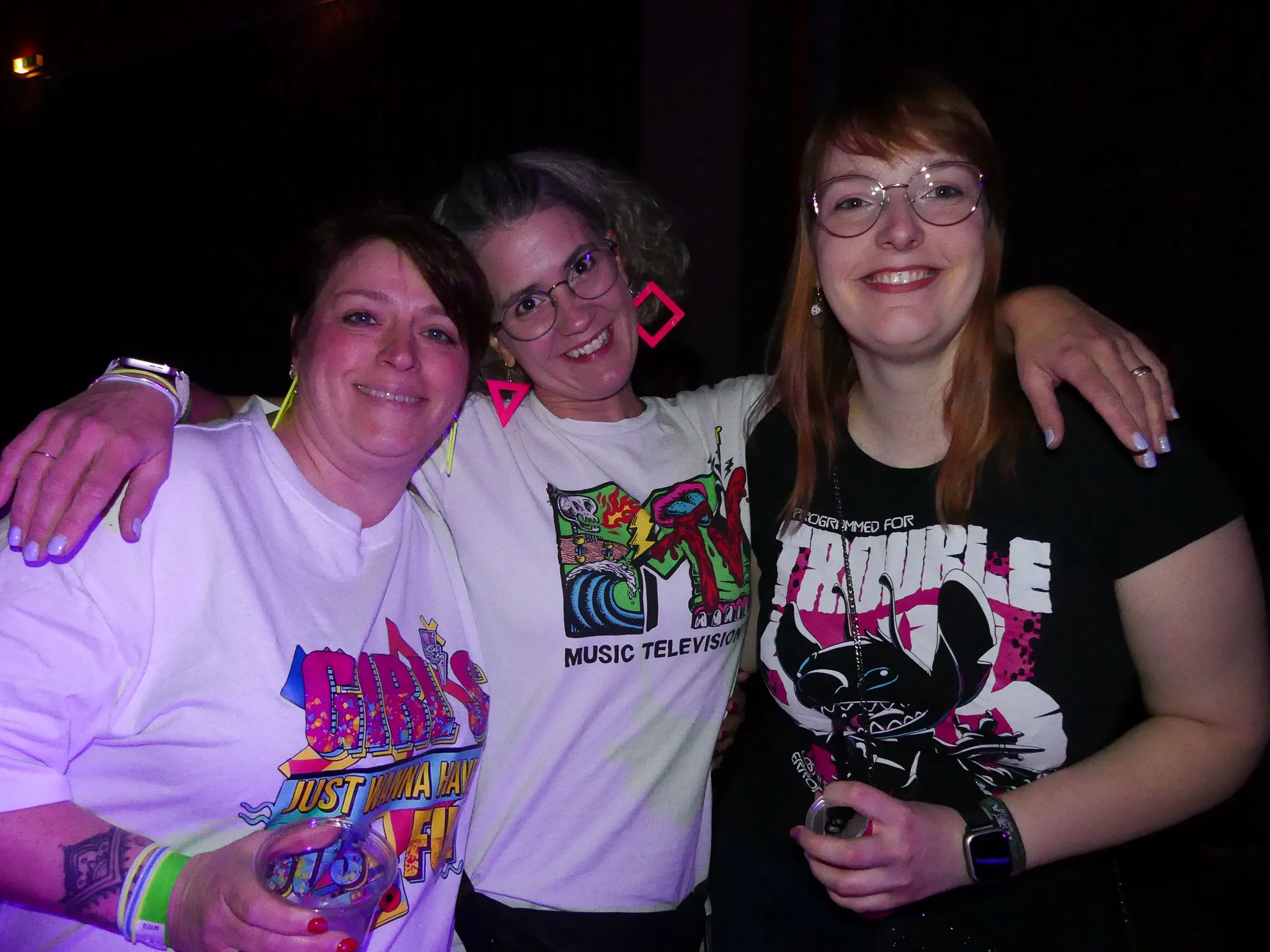 Drei lächelnde Frauen umarmen sich auf der Tanzfläche einer 80er-/90er-Party. Sie tragen farbenfrohe T-Shirts mit Motiven aus dieser Zeit.