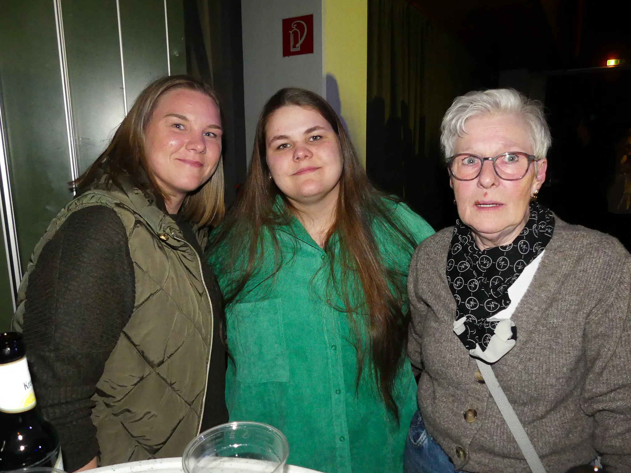 Drei Frauen unterschiedlichen Alters posieren eng nebeneinander. Die jüngste trägt eine Weste, die mittlere ein grünes Hemd und die ältere eine graue Jacke mit Brille.